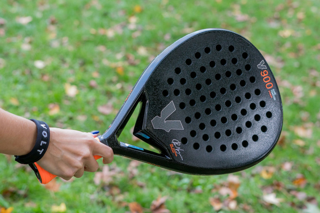 A hand holding a padel racket with grass in the background.